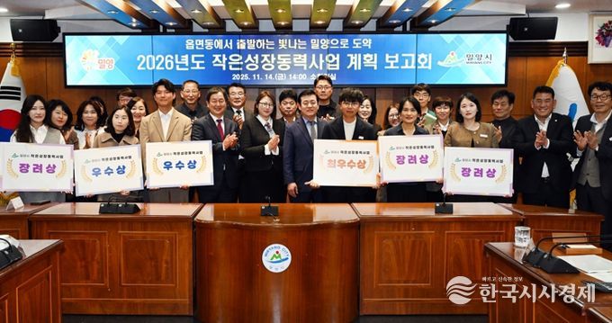 밀양시청 소회의실에서 열린 2026년도 작은성장동력사업 계획 보고회에서, 평가 결과 선정된 읍·면·동 직원들이 사업 선정 표찰을 들고 단체 사진을 촬영하고 있다.